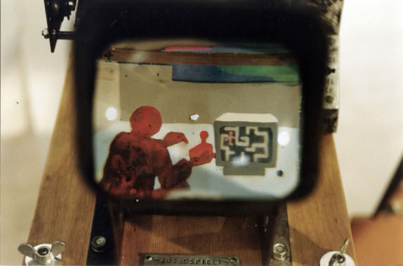 Einblick in das Mutoskop 2 "ausgespielt" von Basil Vogt. Eine Figur spielt am TV ein Videospiel (Vorläufer des Computerspiels oder Playstation). Die Figur macht eine Pause streckt sich und merkt, dass sie selbst in ihrem Computer eine Figur spielt. Völlig orientierungslos irrt die Figur durch das Labyrinth in welchem sie durch welches sie gehetzt wird. Wir steuren und werden gesteuert im endlosen Loop und im Rückzoom. Die Bilder sind von zwei Seiten auf ein angeschliffenes Plexiglas mit Acrylfarben gemalt.